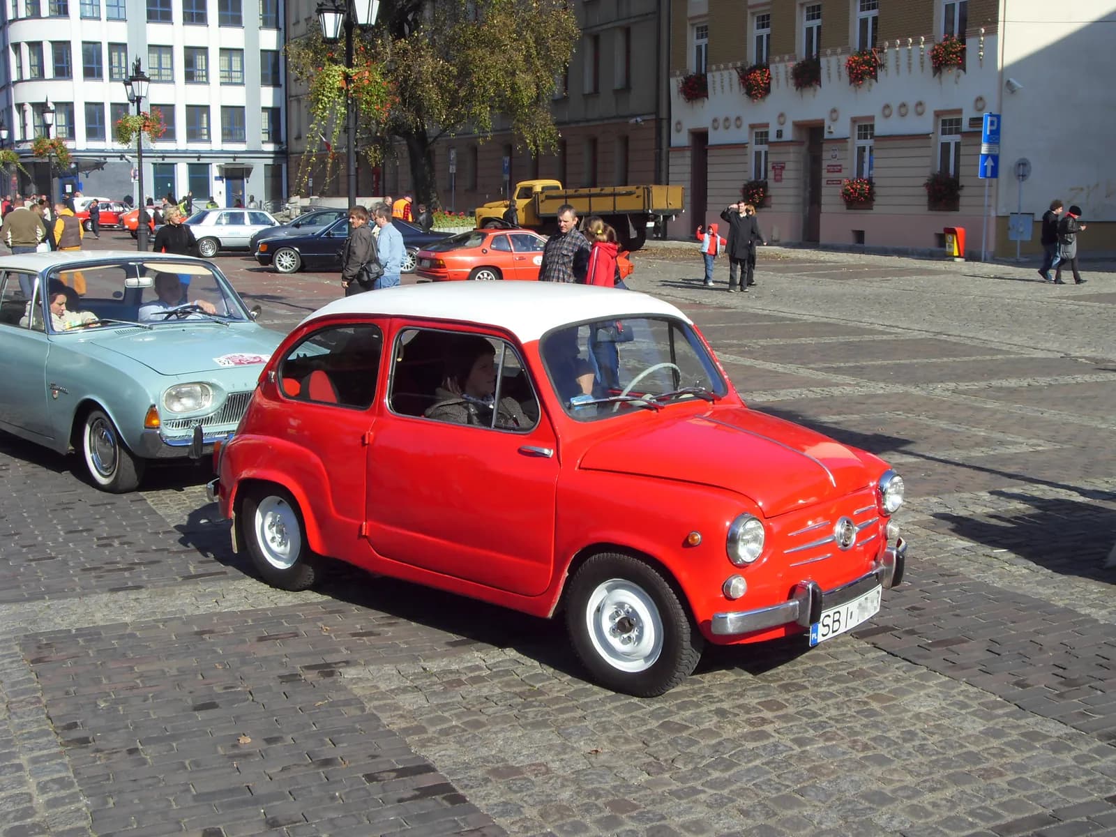 A bright red vintage car with a white roof is parked on a cobblestone street, with another light blue vintage car partially visible behind it, and people and buildings in the background.