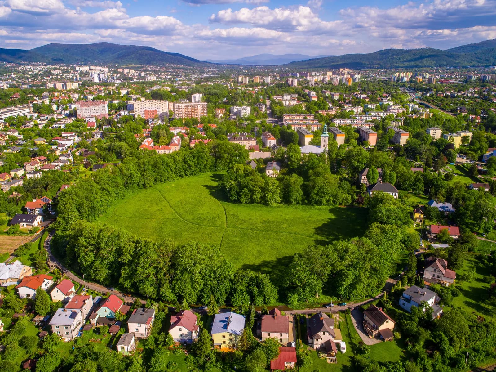 An aerial view shows a sprawling town with a circular green park at its center, surrounded by buildings and distant mountains under a blue sky.